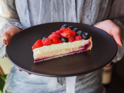 Person Holding a Black Plate with a Slice of Cheesecake with Fresh Berries