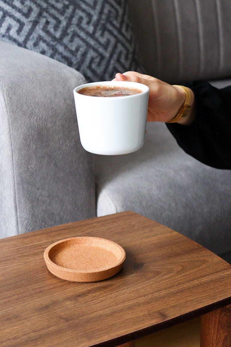 A Person Holding A Ceramic Mug Near The Wooden Table With Brown Coaster