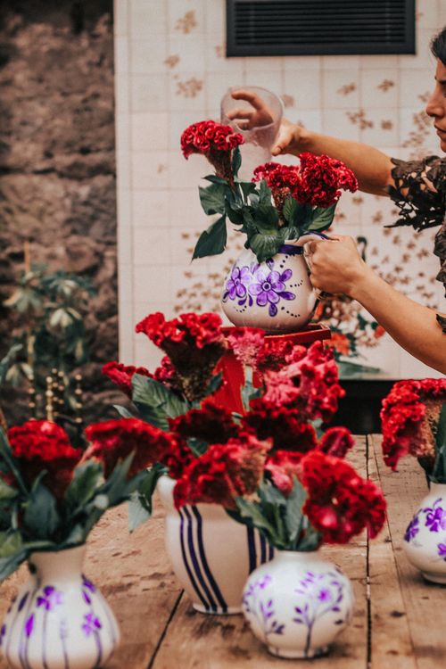 Free A woman artfully arranges vibrant red flowers in decorative vases on a wooden table. Stock Photo