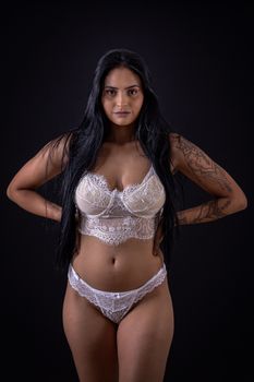Stunning portrait of a woman posing confidently in white lingerie, showcasing her tattoos against a black background.