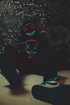 A person wearing a neon LED mask sits in a dark room with graffiti-covered walls.
