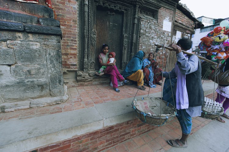 Person Selling Eggs Talking To Women Sitting By House Entrance