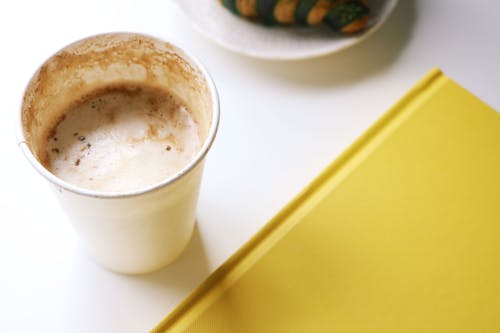 From above closeup of started takeaway cappuccino placed on white table near yellow notebook and plate with croissant