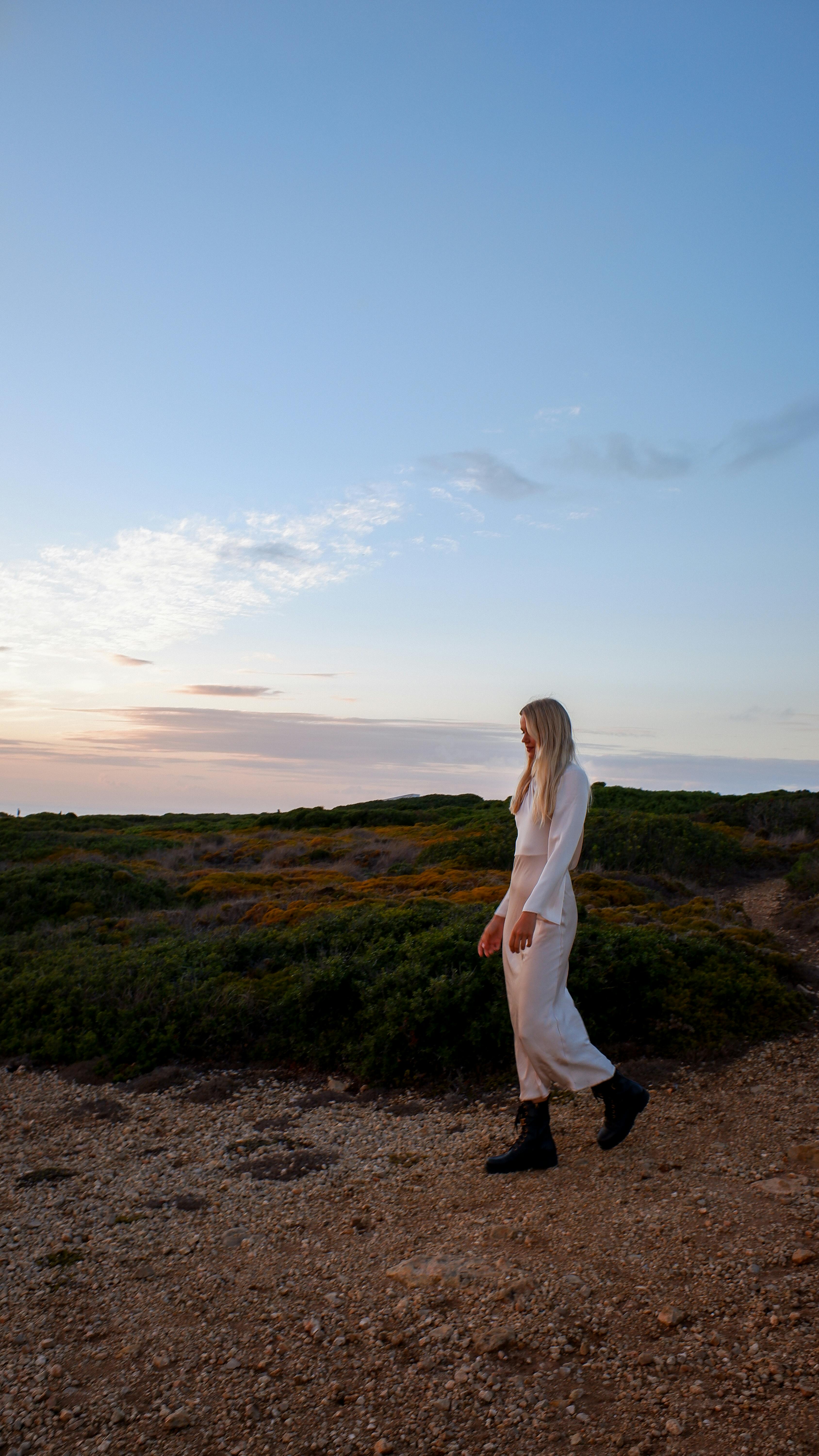 A woman in white clothing walks on a dirt path during a beautiful sunset outdoors.