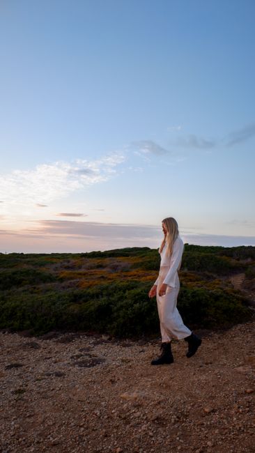A woman in white clothing walks on a dirt path during a beautiful sunset outdoors.