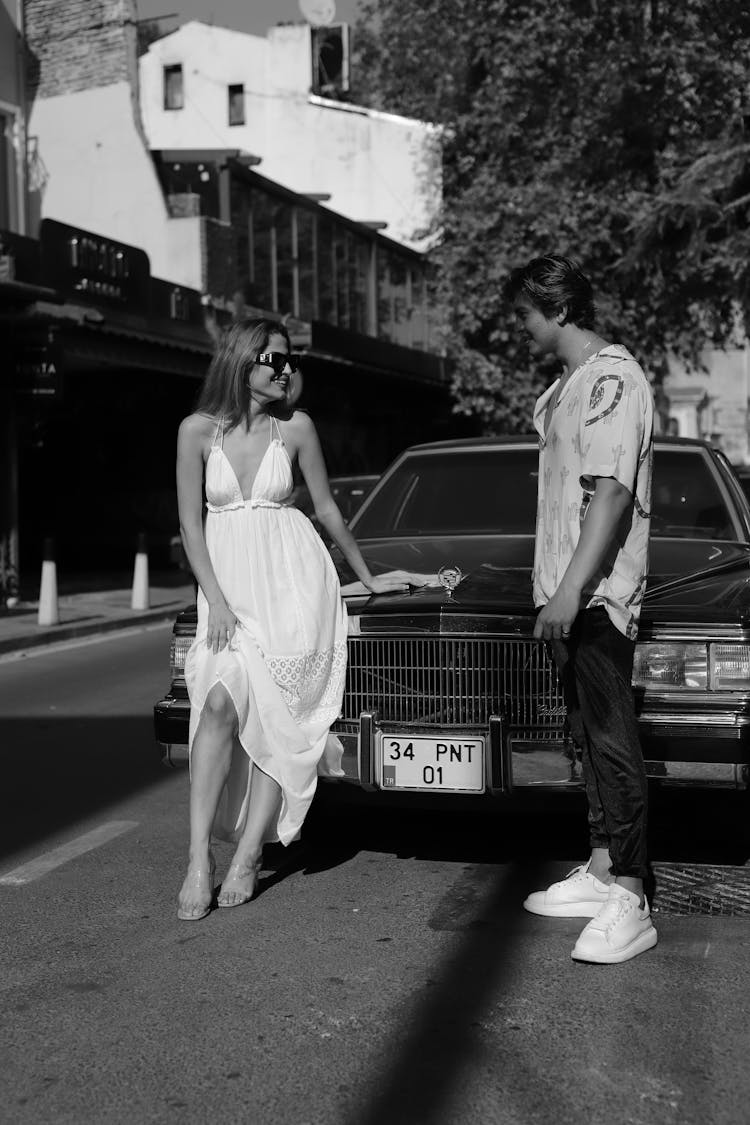 Man Standing Near A Woman Sitting On A Car's Hood