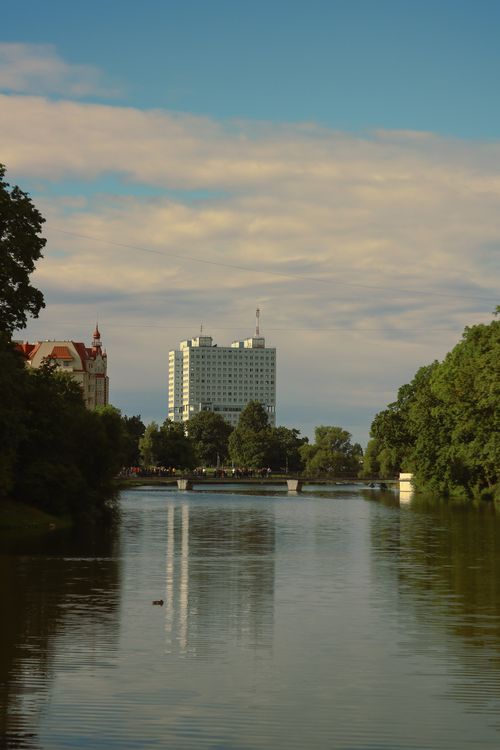 Free Calm river reflecting a cityscape with green trees and blue sky overhead. Stock Photo