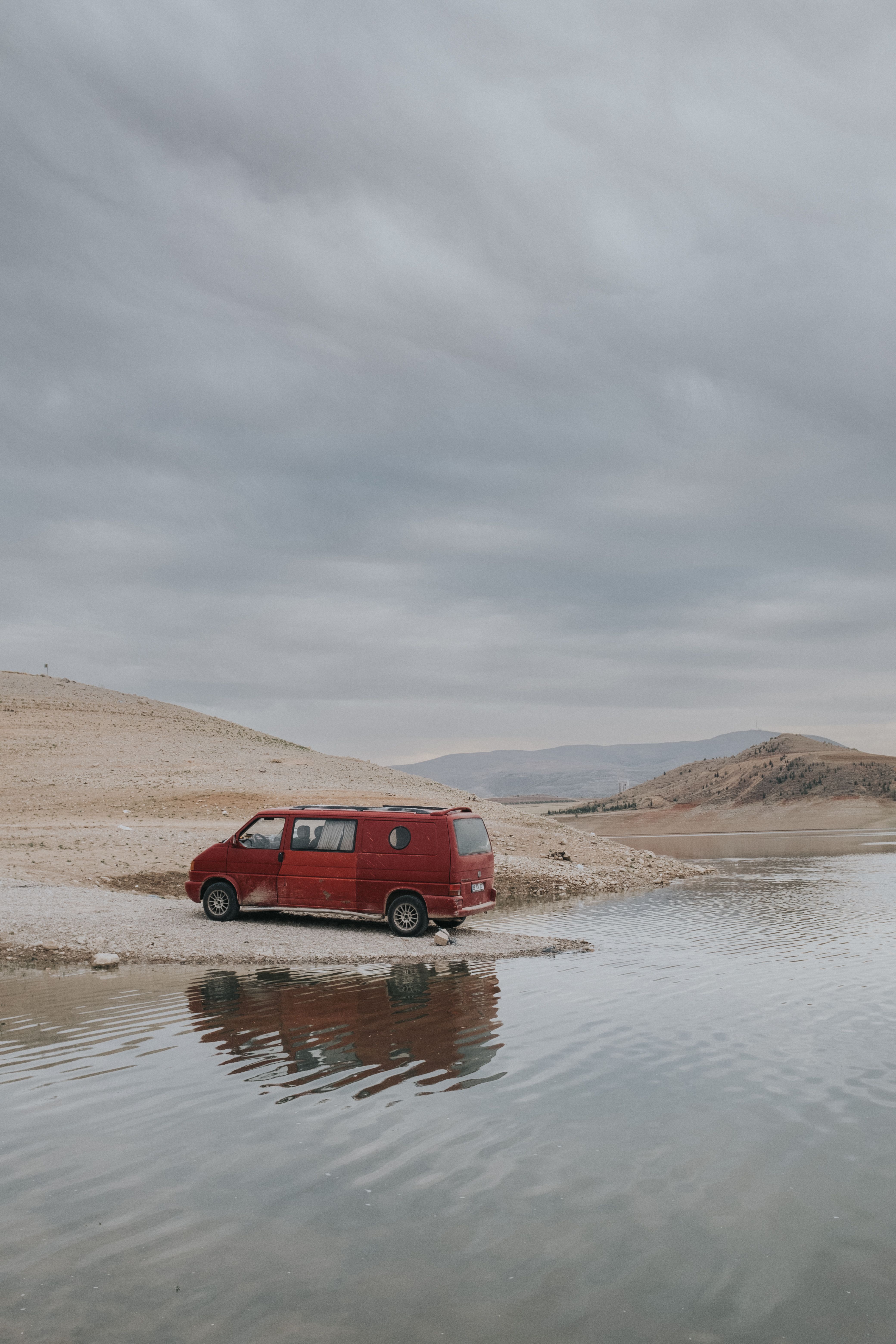 Red van parked on a serene riverbank under a cloudy sky, reflecting in the water.