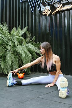 A woman performs a seated stretch in a gym setting with a kettlebell nearby, highlighting fitness and flexibility.