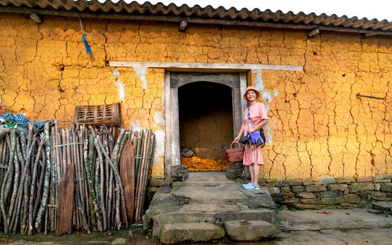 A smiling woman stands at the entrance of a rustic, cracked wall house in a village setting, holding a basket.