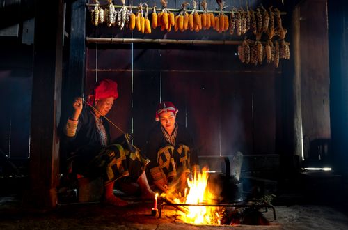 Free Two women in traditional attire cook by an open fire inside a rustic kitchen with hanging corn. Stock Photo