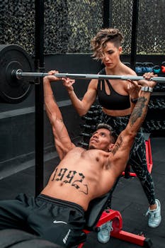 A focused young man lifting weights while a woman assists at the gym.