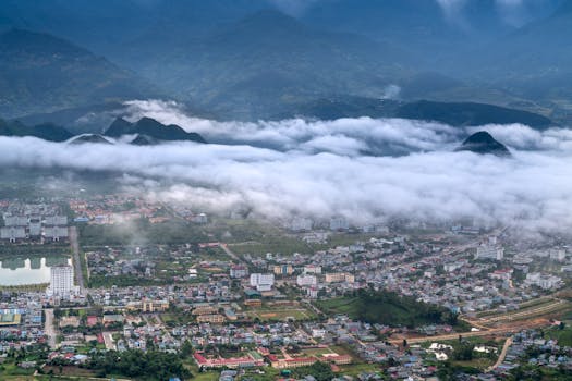 Aerial city view with buildings under a blanket of clouds, surrounded by mountains.