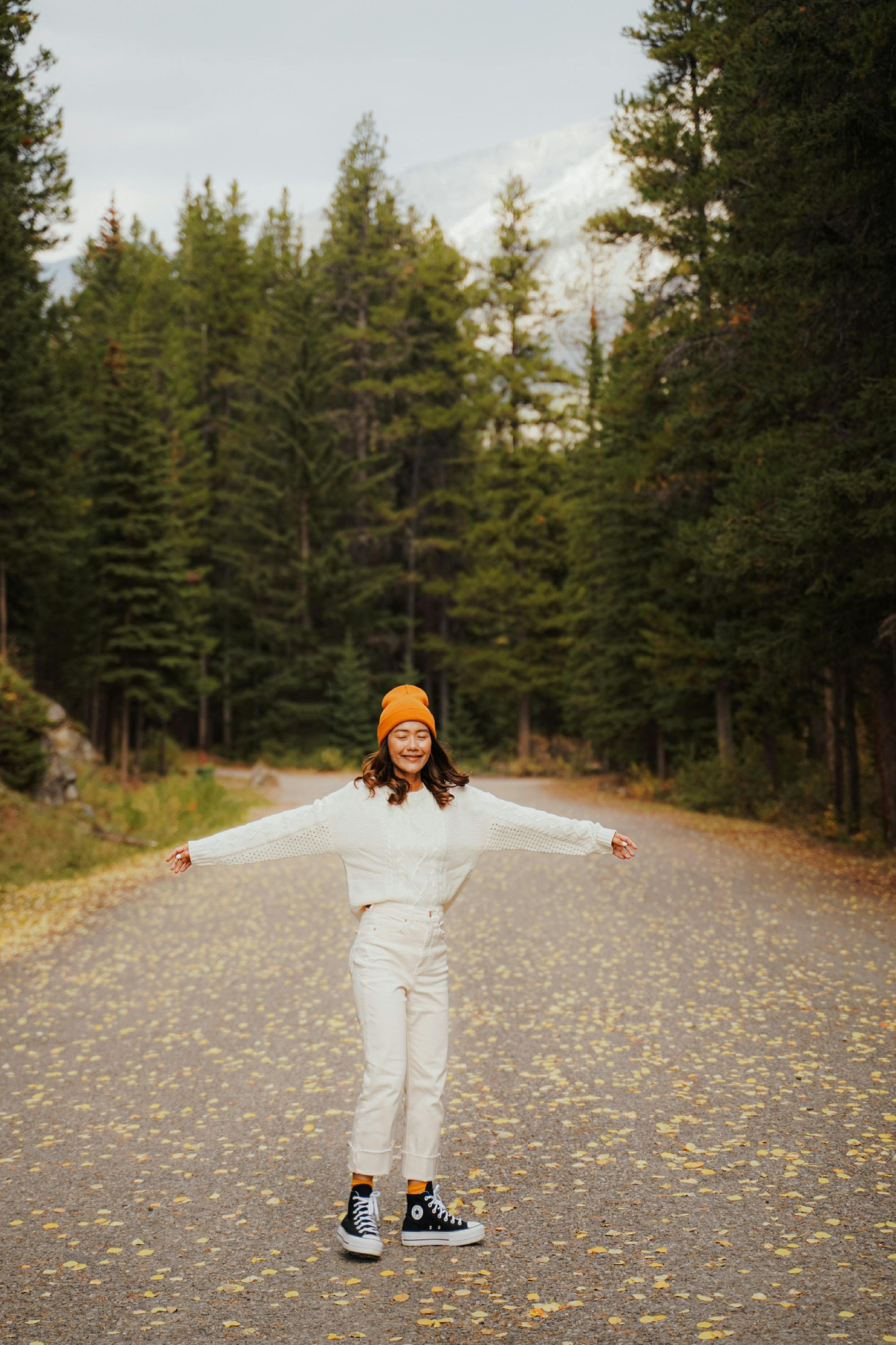 Woman in stylish fall fashion with yellow beanie, posing on a forest road.