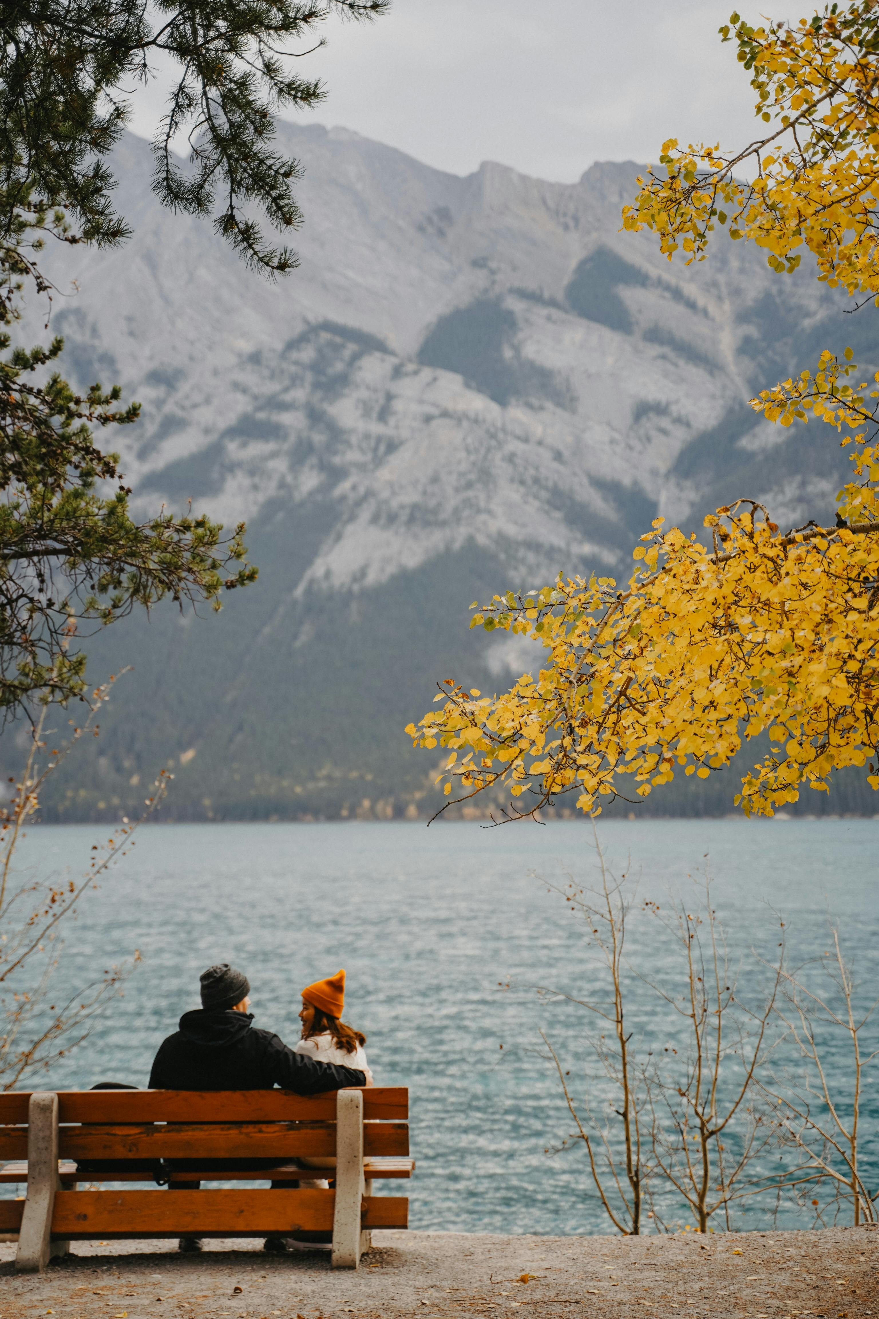 A couple sits on a bench by a lake with yellow leaves and mountains in the background.