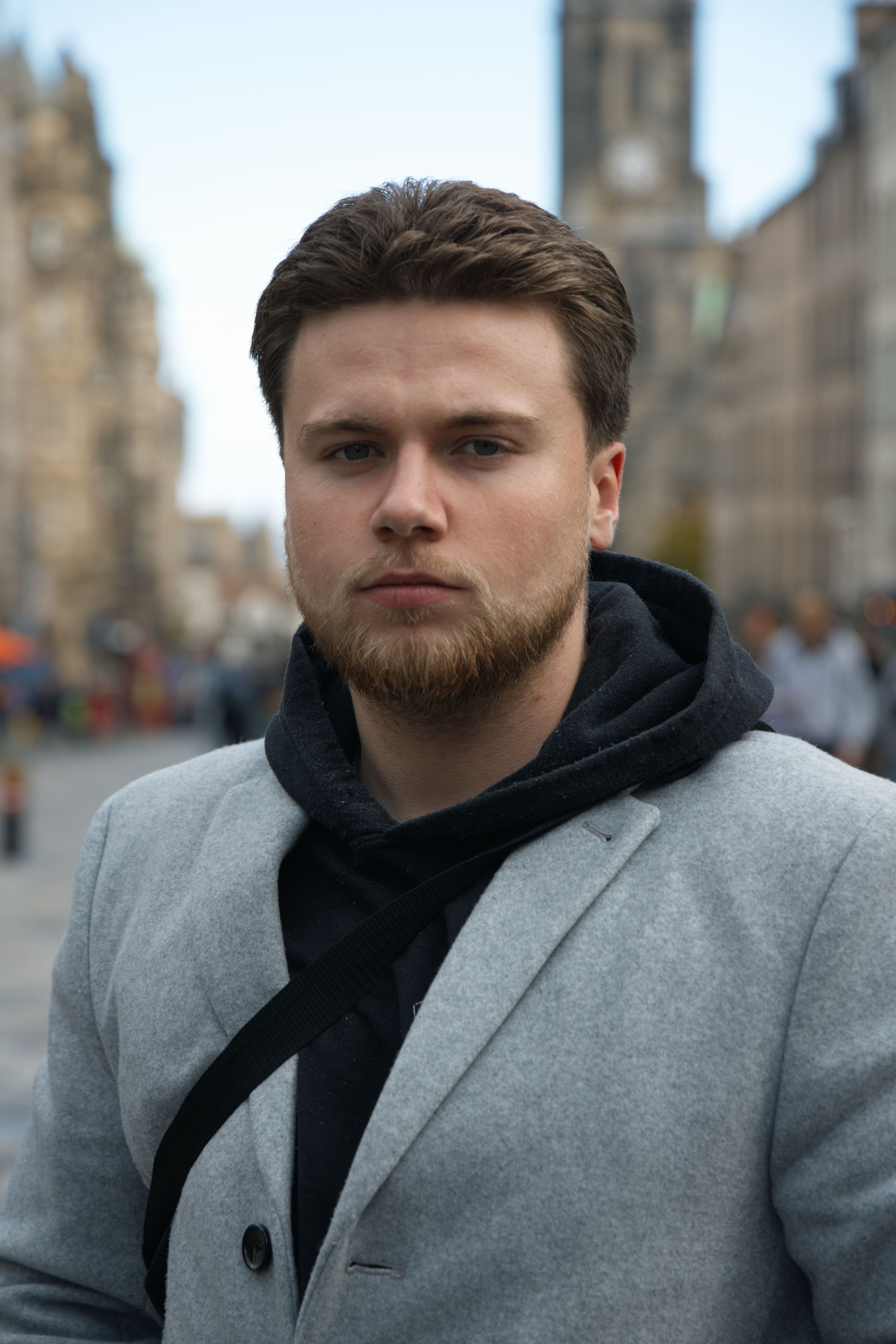 Close-up portrait of a bearded man in a grey coat and hoodie in an urban setting.