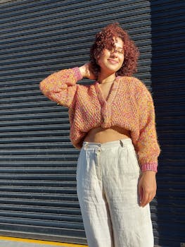 Portrait of a smiling woman with curly red hair wearing a pink sweater in bright sunlight.