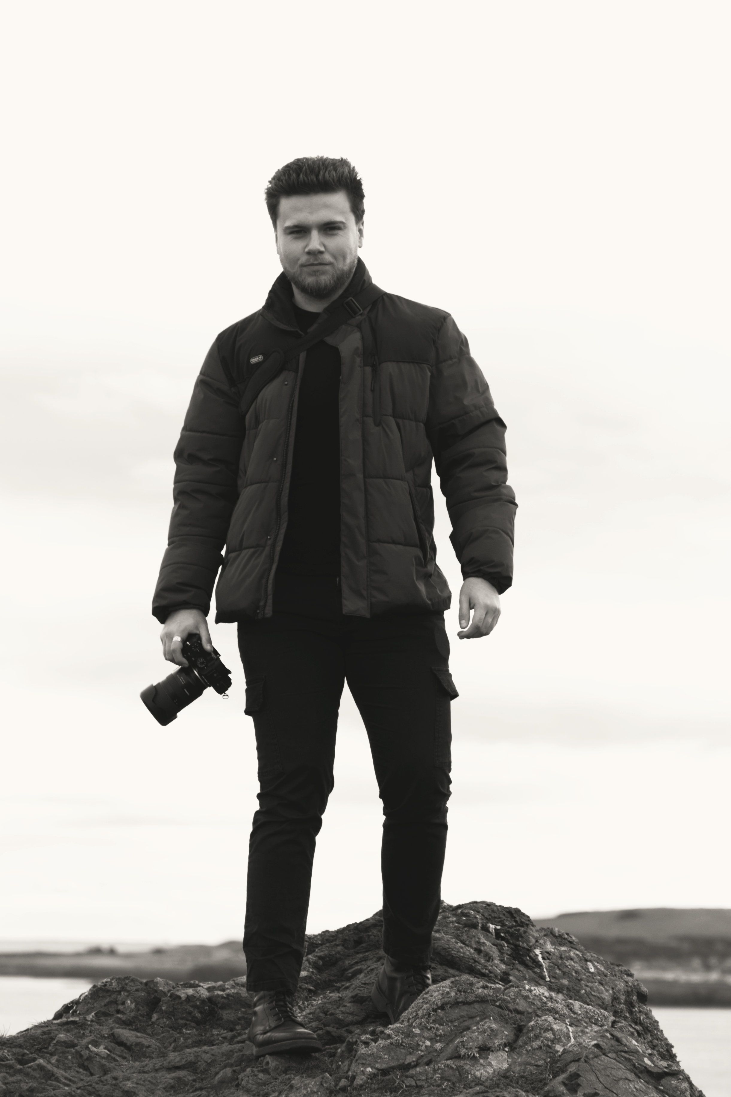 A monochrome image showcasing a photographer with a camera standing confidently on a rocky outcrop outdoors.