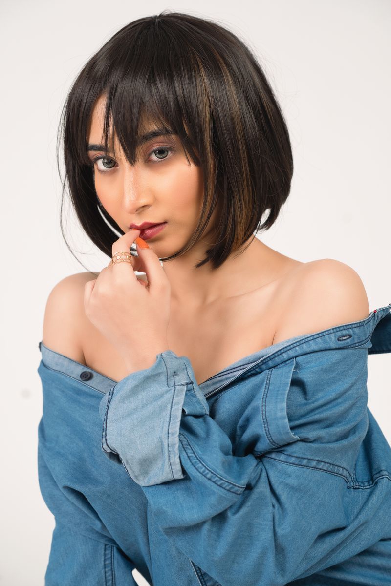 Asian woman with short hair poses in a denim jacket, hand on lips, playful expression.