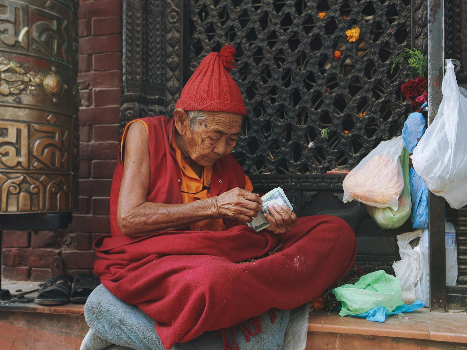 An elderly monk sits cross-legged on a street, counting money amidst bags and traditional Tibetan attire.