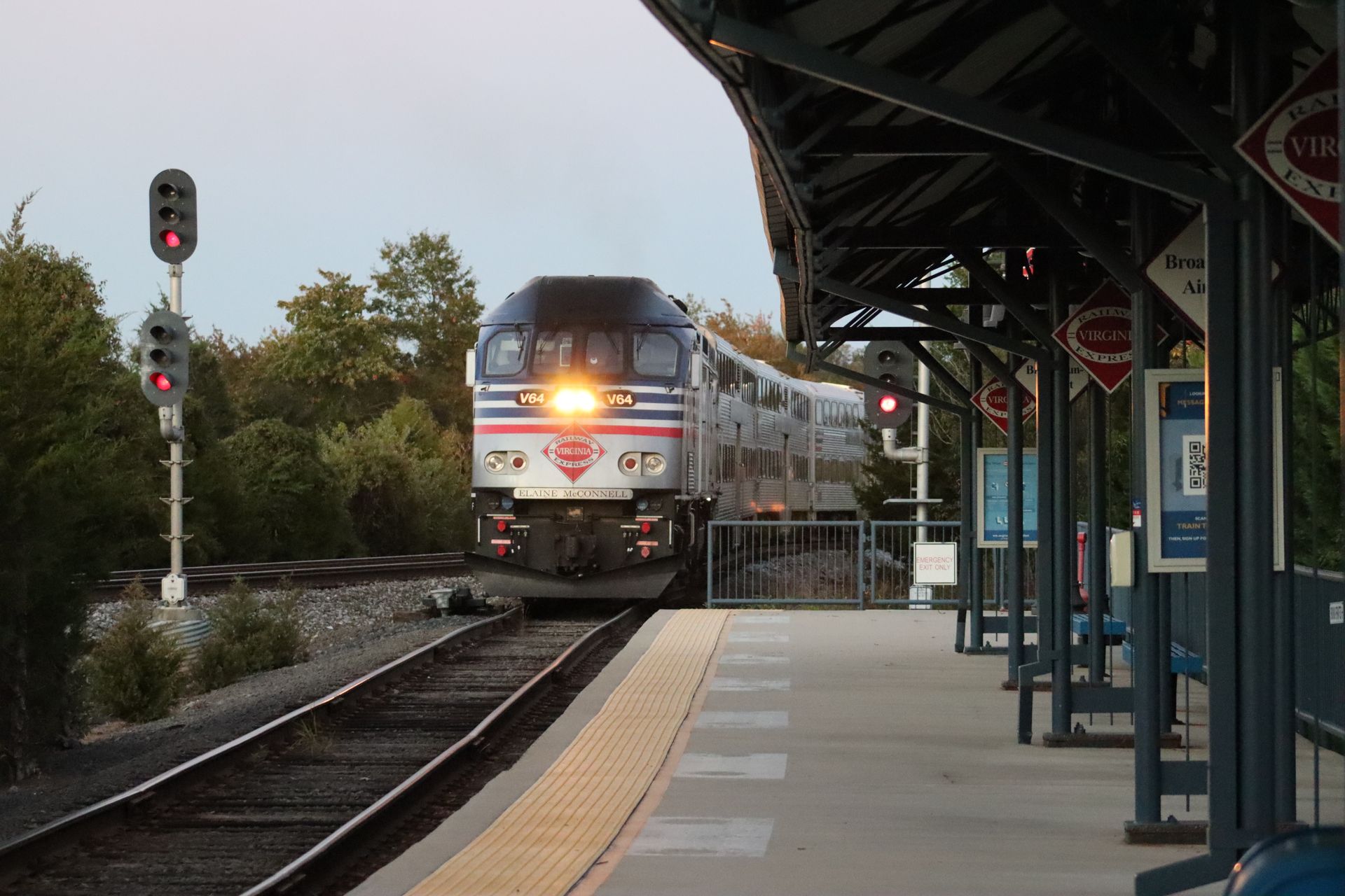 A passenger train approaches a quiet railway platform in daylight, featuring signals and signage.