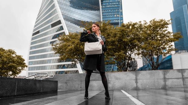 A fashionable woman poses with a bag against a modern cityscape backdrop featuring striking architecture.