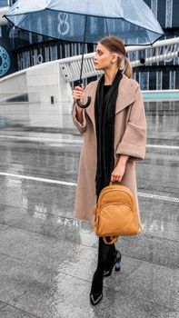 Stylish woman with umbrella and backpack on rainy city street.