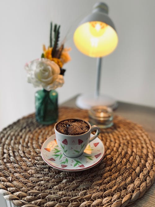 Free A Cup of Hot Coffee on a Saucer Near Desk Lamp and Glass with Flowers Stock Photo