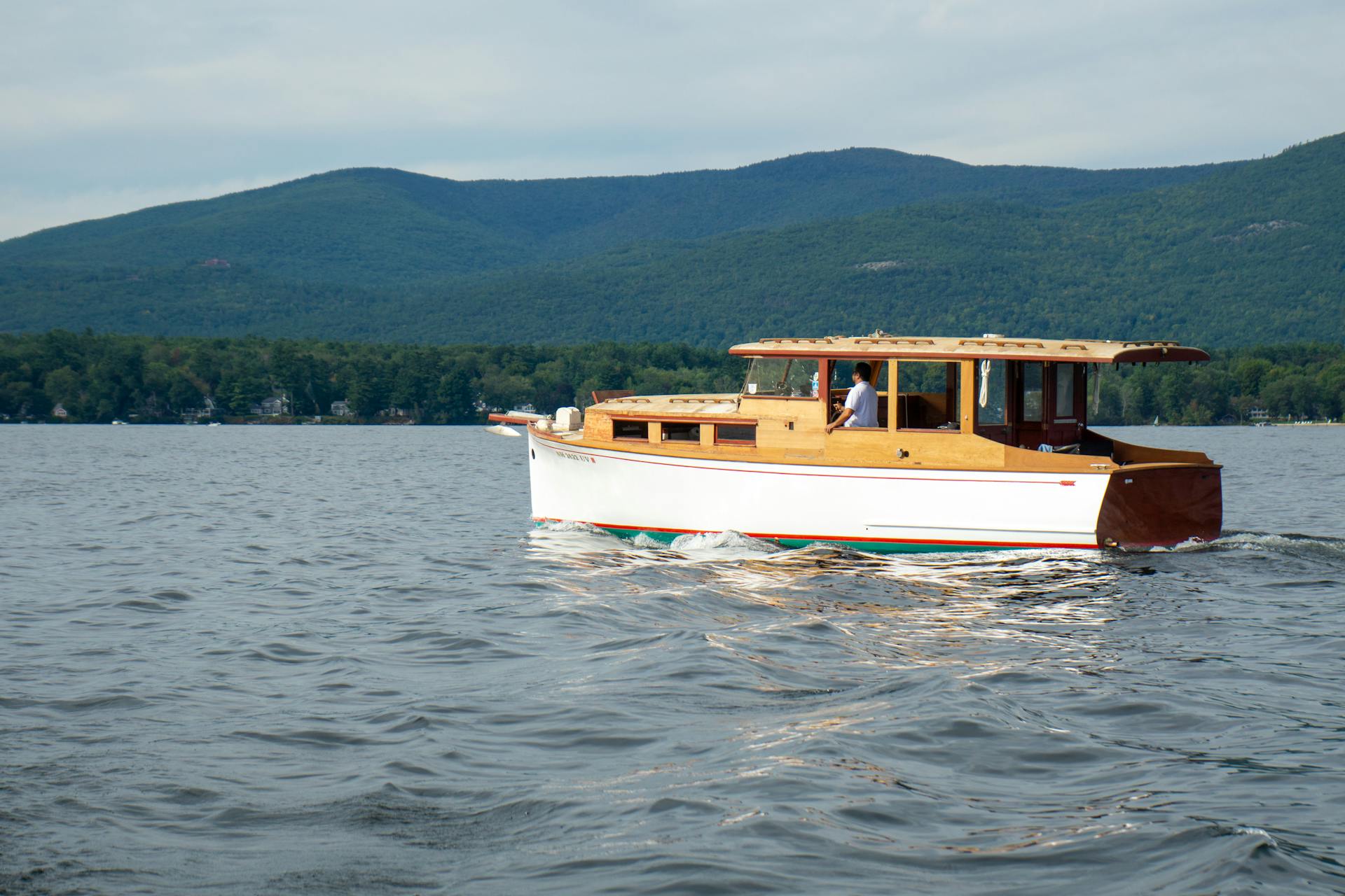 A vintage wooden boat cruises on a calm lake against a backdrop of lush green hills.