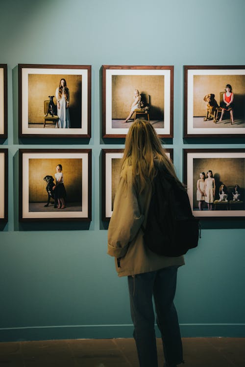 Woman in Brown Coat Standing Carrying Backpack while Looking at the Picture Frames Hanging on the Wall