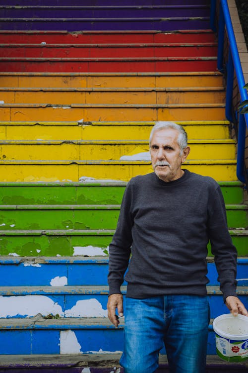 An Elderly Man in Gray Long Sleeves Standing Near Colorful Stairs while Looking at the Camera