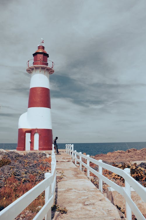 A Person Leaning on the Wooden Railing Near White and Red Lighthouse Under Cloudy Sky