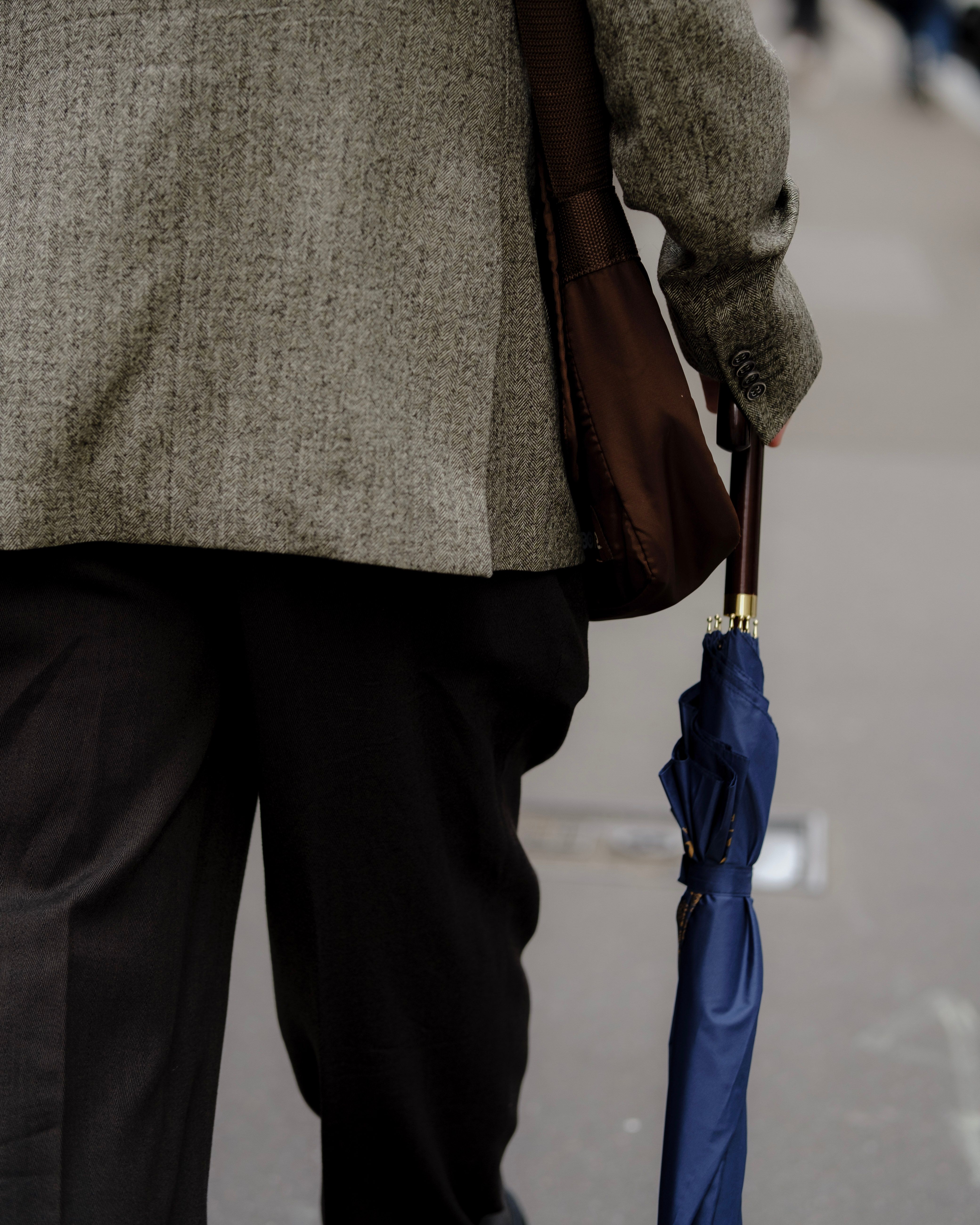 A rear view of a man wearing a blazer, carrying a bag and umbrella, walking outdoors.