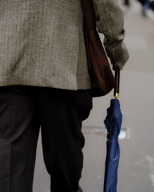 A rear view of a man wearing a blazer, carrying a bag and umbrella, walking outdoors.