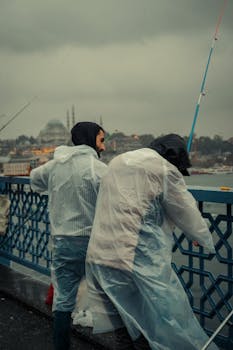Two men in raincoats fishing on a city bridge during a rainy day.