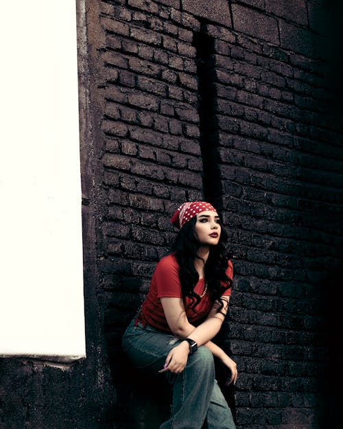 Woman in Red Shirt and Blue Denim Jeans Sitting on Black Concrete Wall