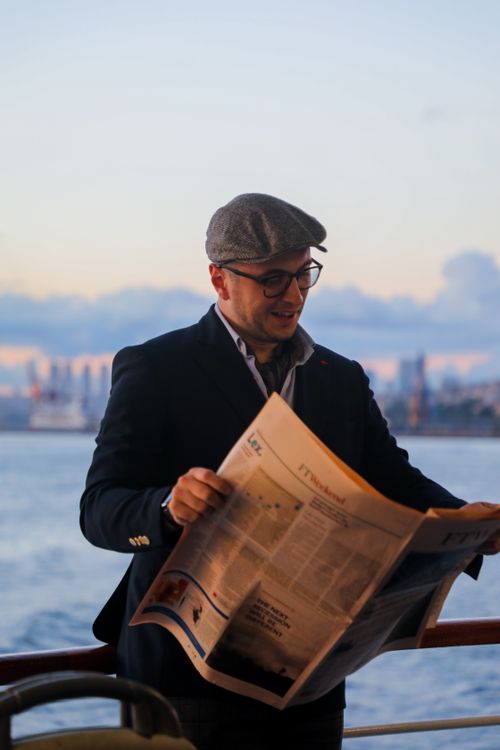 Free Casually dressed man reading a newspaper on a boat with city skyline backdrop at sunset. Stock Photo