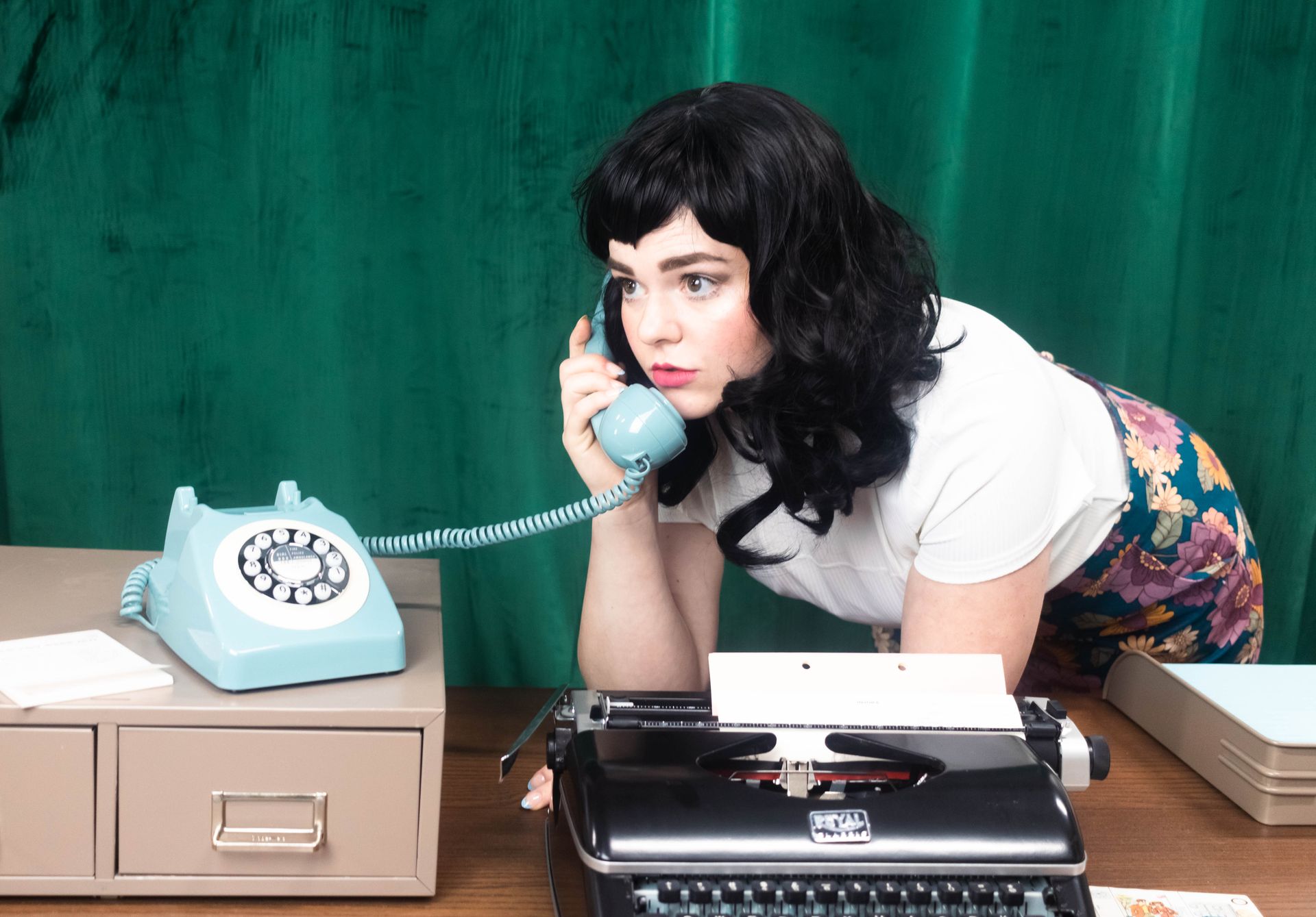Stylish woman in retro office setting using rotary phone and typewriter.