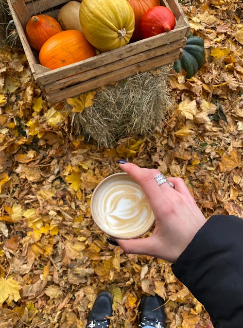 Person Holding a Cup of Coffee Beside a Crate of Squashes