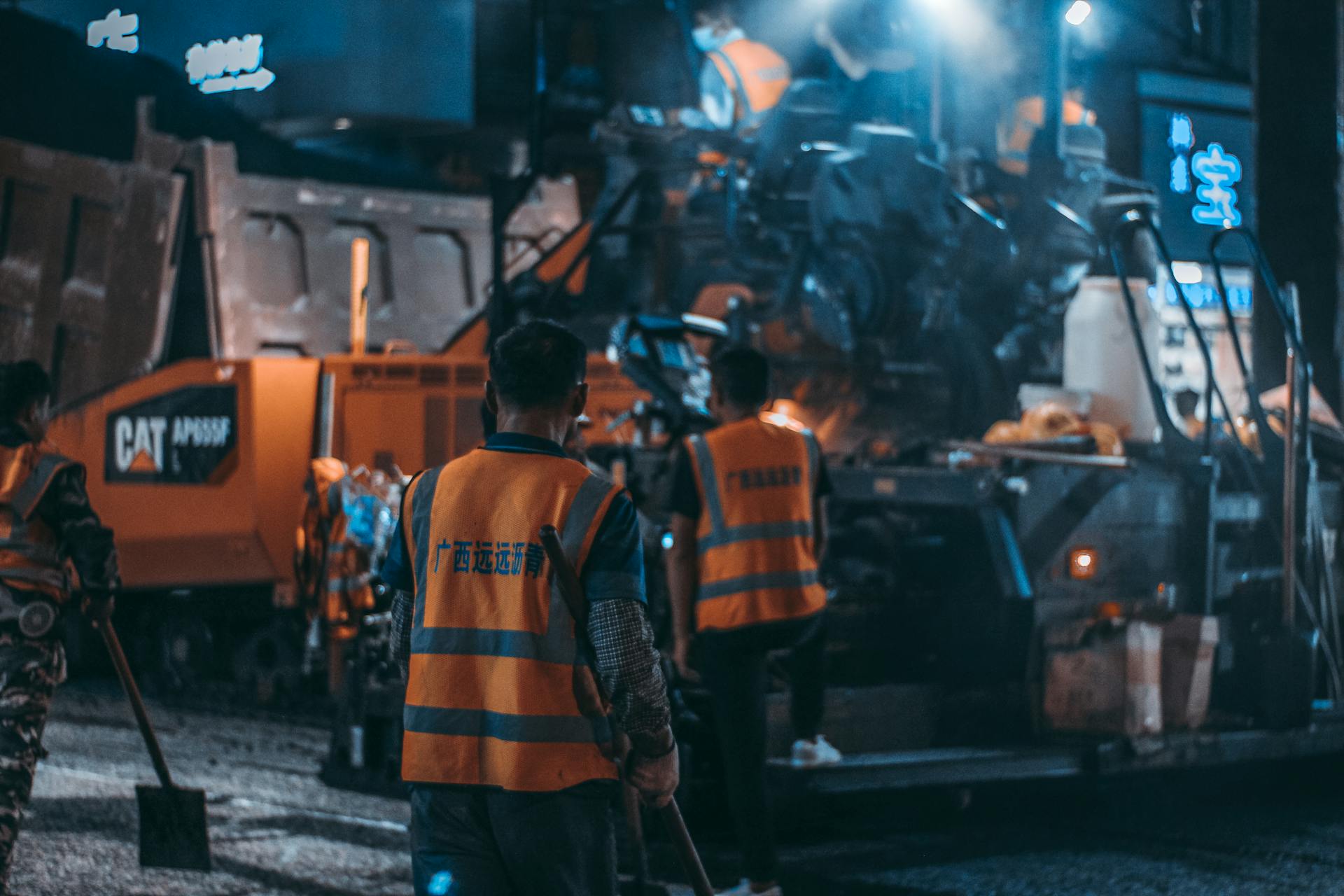 Workers in safety vests operate heavy machinery at a night construction site.