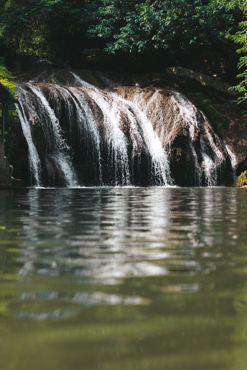 Free A tranquil waterfall flowing over rocks into a reflective pool surrounded by lush greenery. Stock Photo