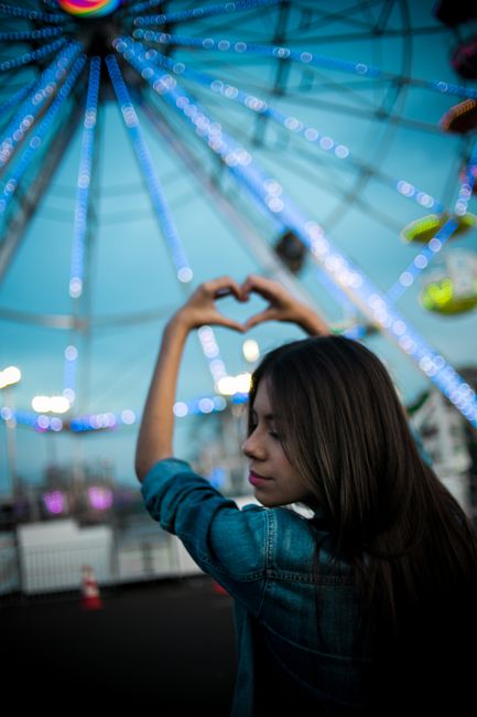 A young woman forming a heart with her hands in front of a brightly lit Ferris wheel at a night festival.
