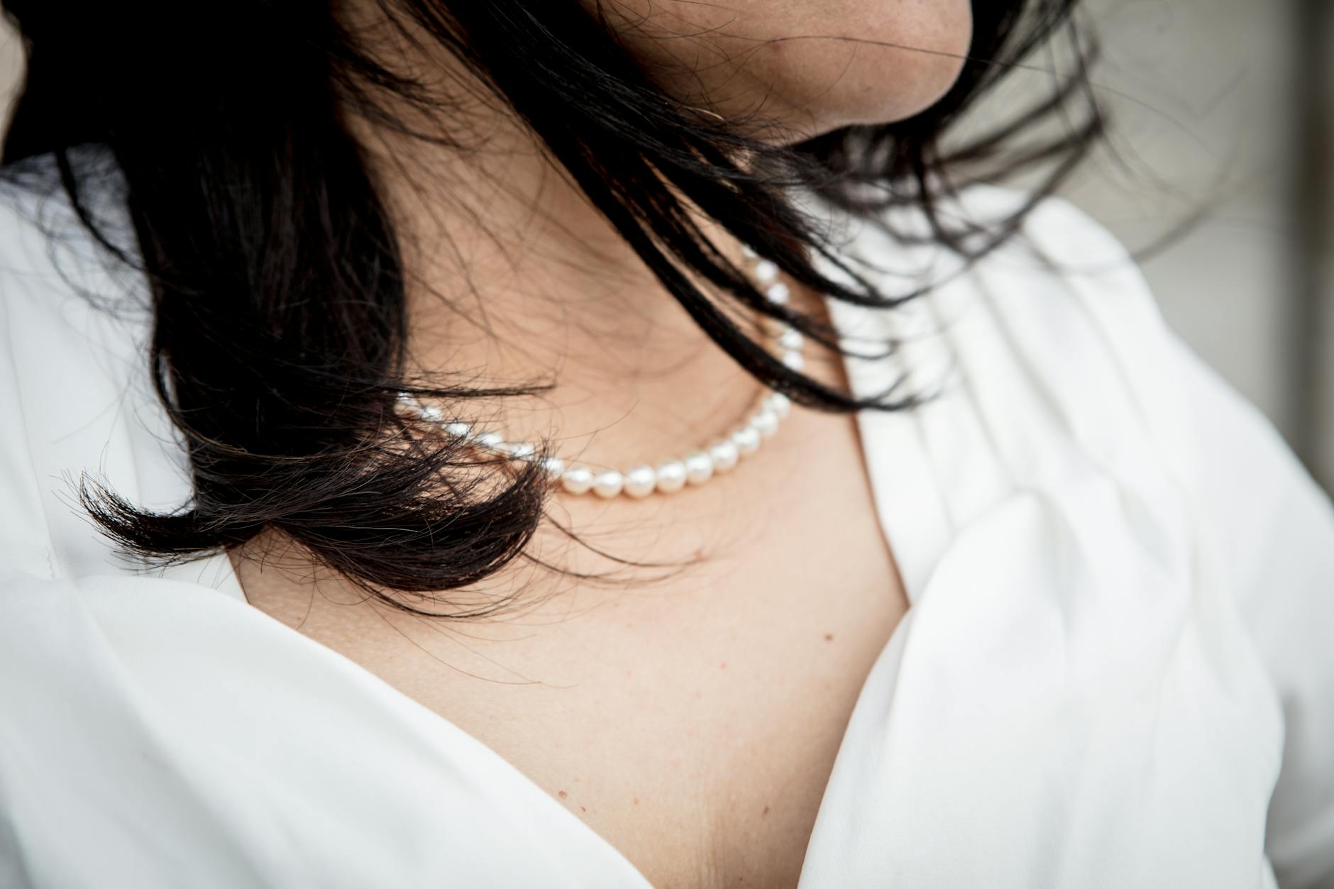 A close-up view of a woman wearing an elegant pearl necklace, complemented by her flowing dark hair and white blouse.