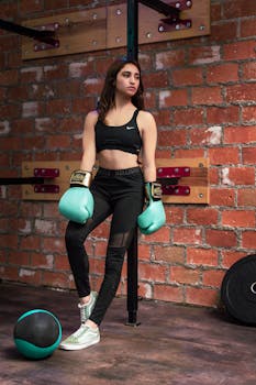 Confident female athlete wearing boxing gloves leaning against a gym wall.