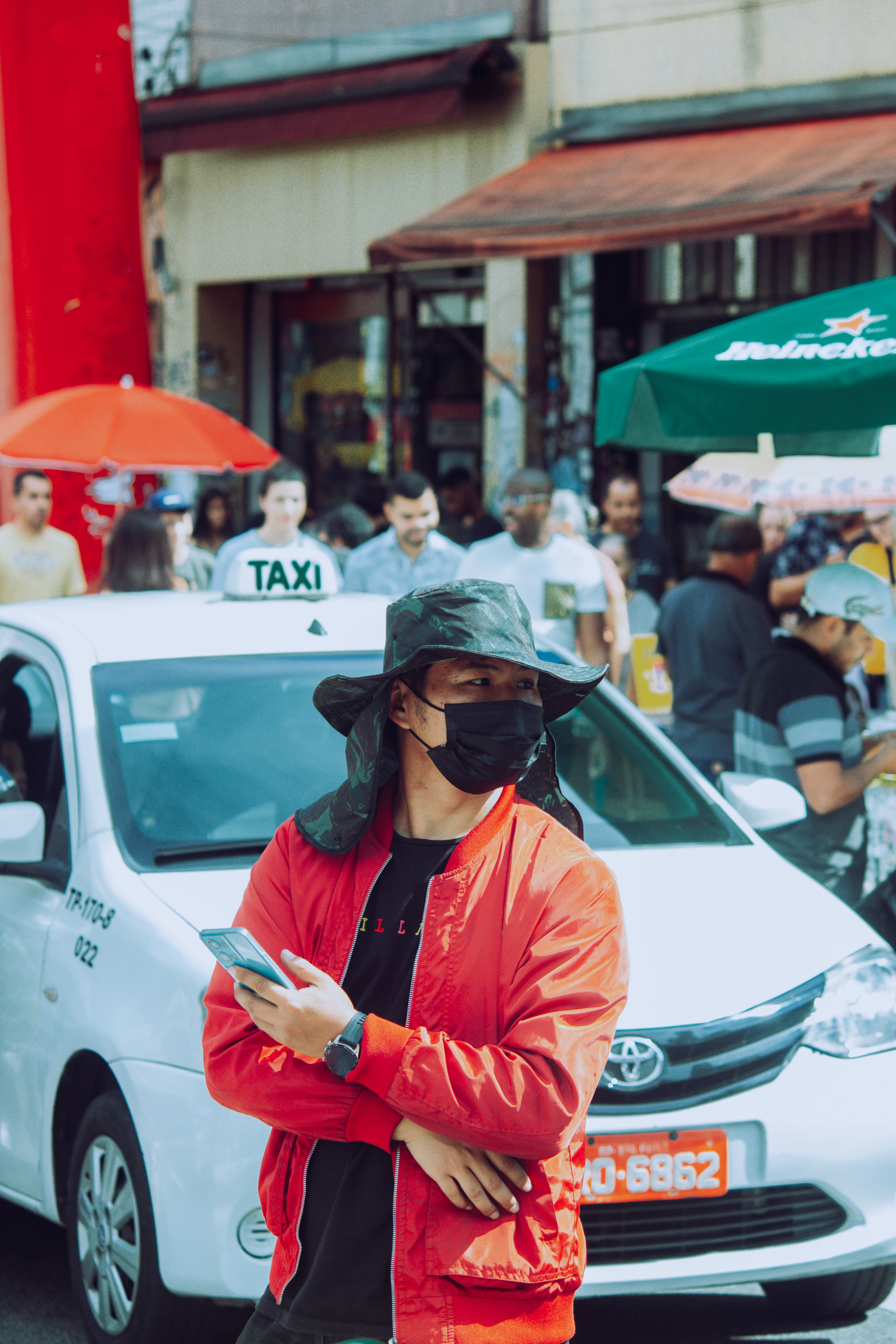 Man with face mask and hat stands by taxi in busy city street.