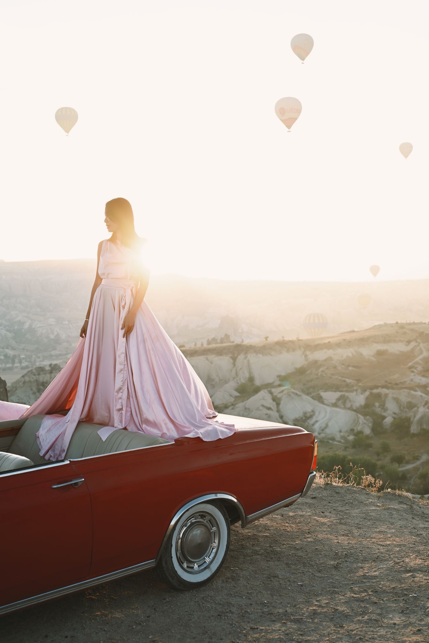 A woman in a dress on a retro car in the mountains at sunset — pink dressing, golden light