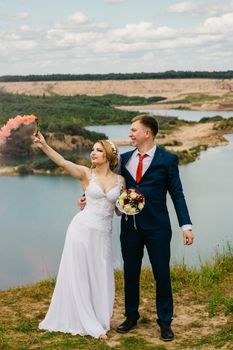 A joyful bride and groom celebrate their wedding with a smoke bomb by a scenic lake.