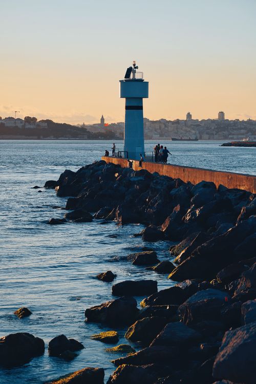 Free A serene lighthouse at dusk with people walking along a rocky pier, overlooking calm waters. Stock Photo