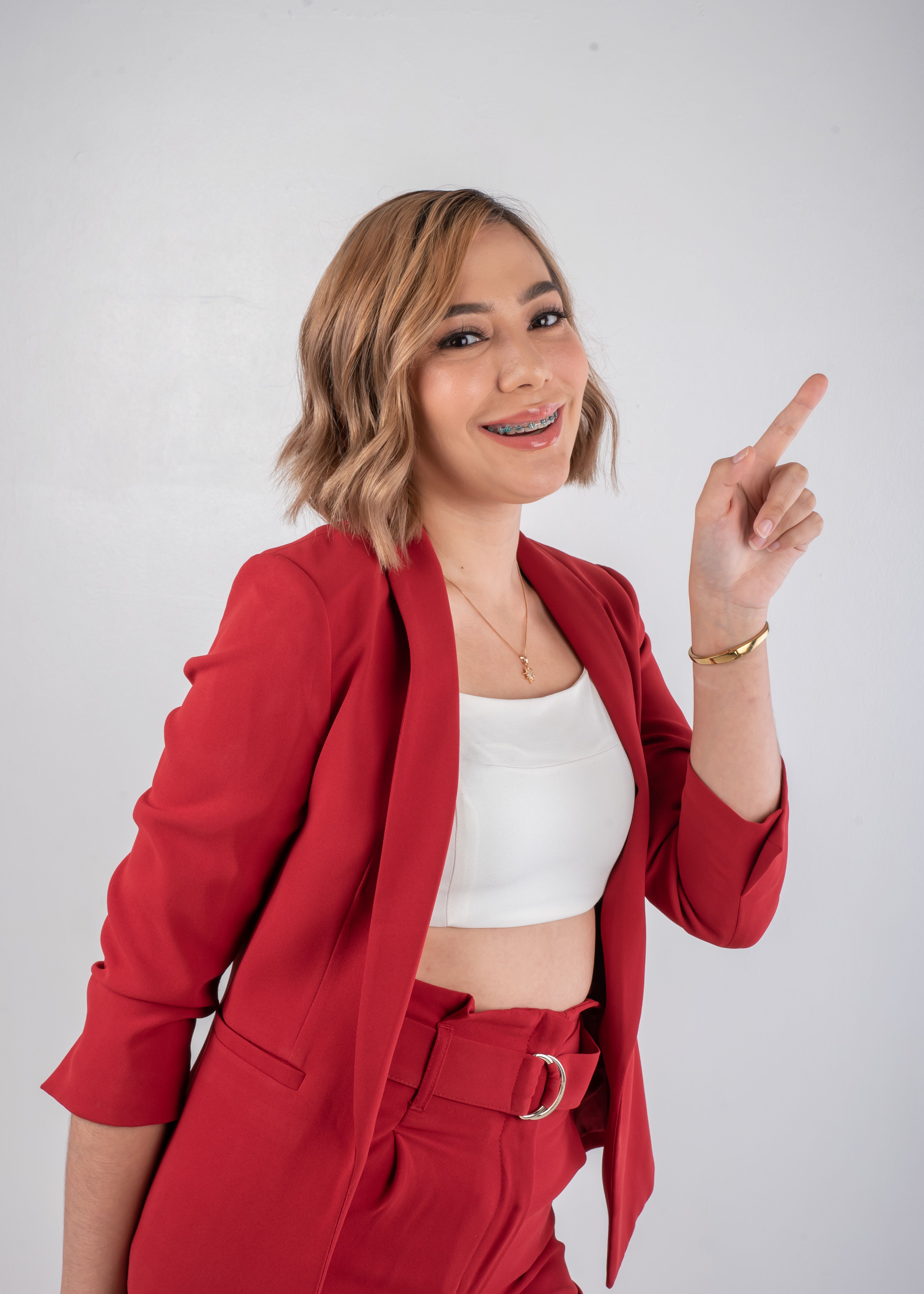 Confident woman in a stylish red suit smiling and pointing up in a professional studio setting.