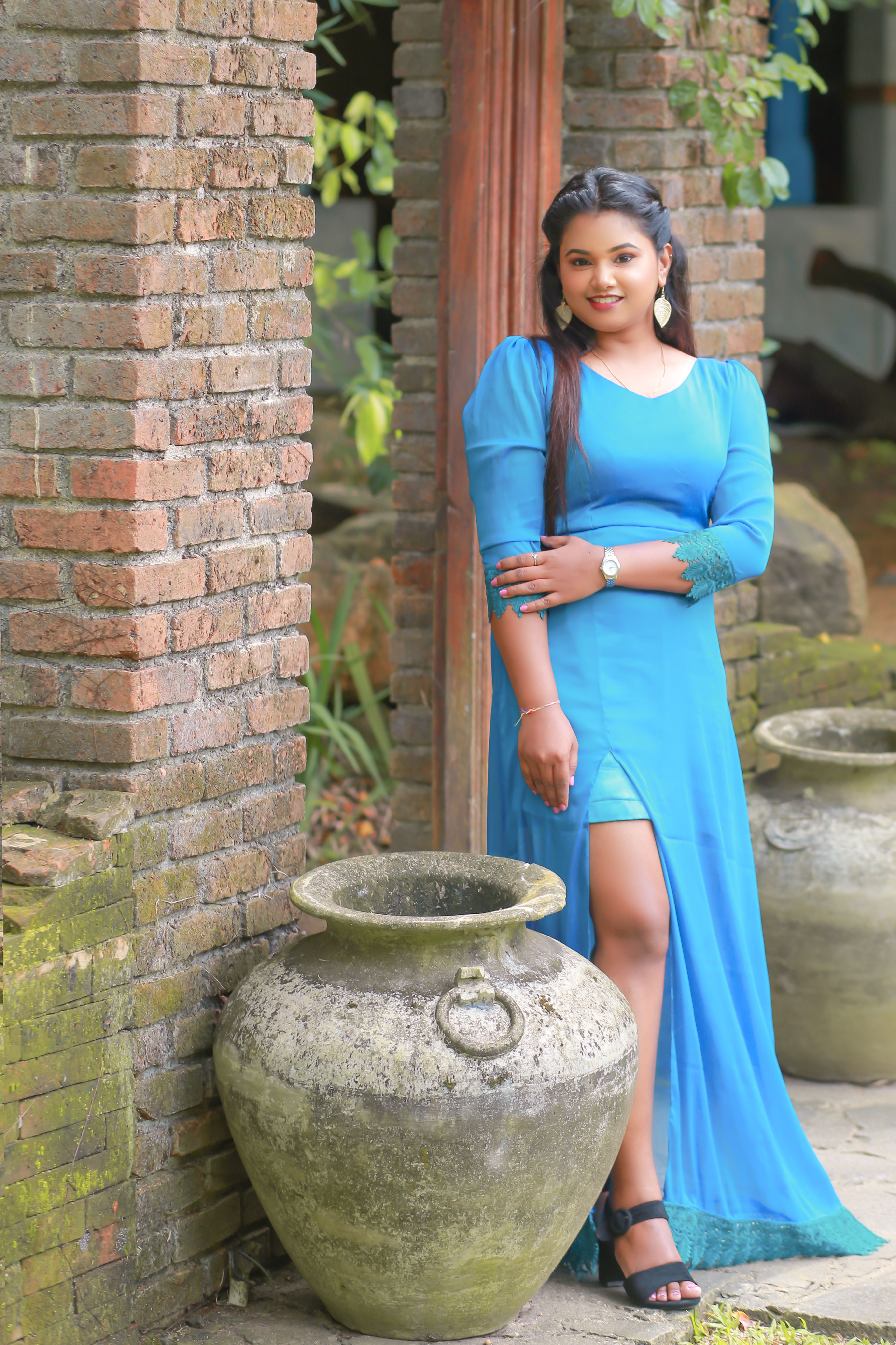 Smiling woman in a blue dress standing by a brick wall and large jar outdoors.